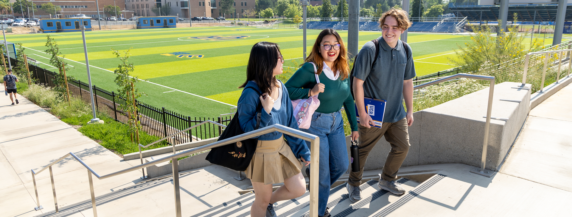 Three students walk up the steps next to the UC Riverside Soccer Stadium and move in the direction toward the Student Recreation Center. There are two female students and one mail student. The soccer field in the background is a vibrant color of green grass.