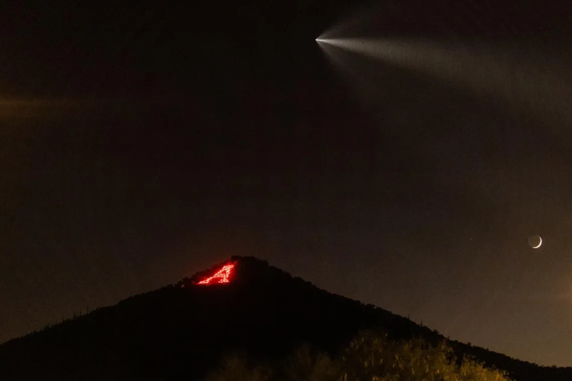 The "A" on Sentinel Peak lit up with flares as a SpaceX rocket flies above