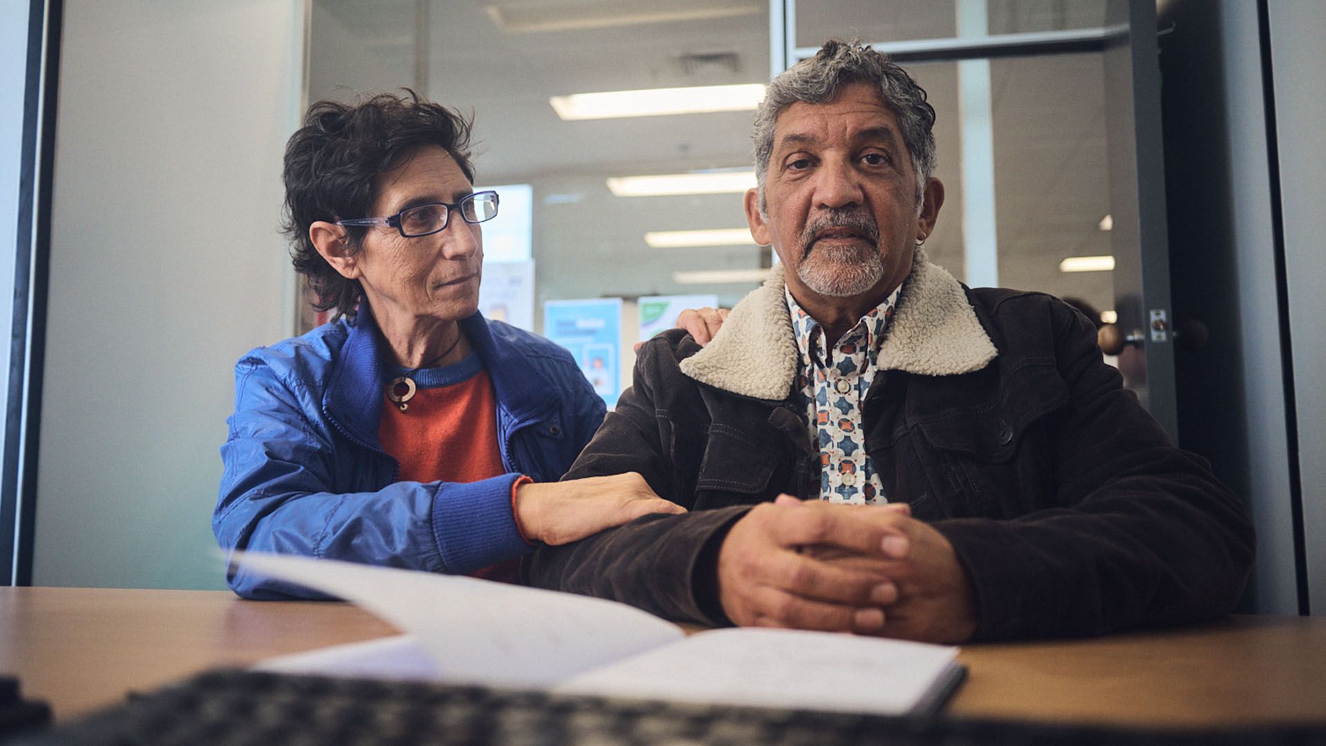 Photo of solemn-looking man receiving serious news at a medical appointment while flanked by his wife.