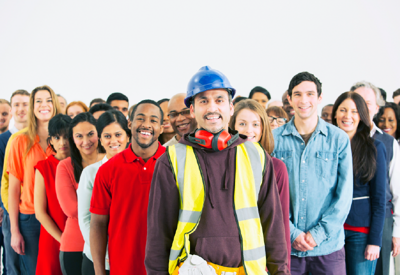 A man wearing a hardhat, ear protection and a high-visibility vest stands in front of a diverse group of smiling workers. 