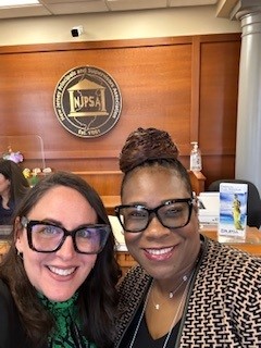 Two women wearing funky black glasses smile for a selfie. The NJPSA logo is on the wall in the background.