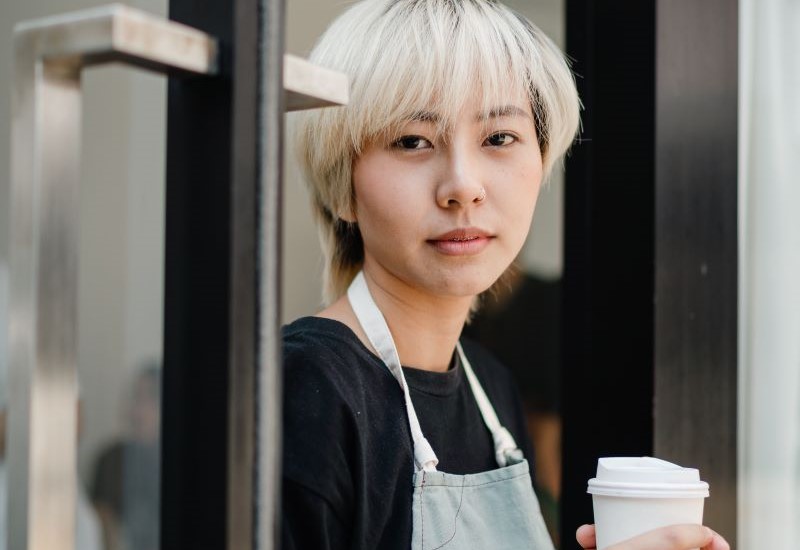 An Asian woman with platinum blond hair holding a cup of coffee wearing a black t-shirt and an apron.