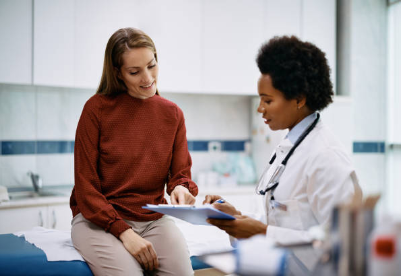 A white woman in a read turtleneck seated in a doctor's office with a Black woman doctor in a white coat and holding a clipboard.