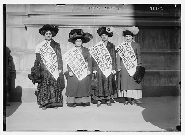 Black and white photo of garment workers striking during the "Uprising of the 20,000" in New York City, February 1910. Four women wearing long layered dresses and large hats are also wearing sashes with "Picket Ladies Tailors Strikers"