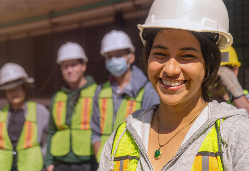 Una joven trabajadora con casco sonríe, mientras otros trabajadores están de pie detrás de ella.