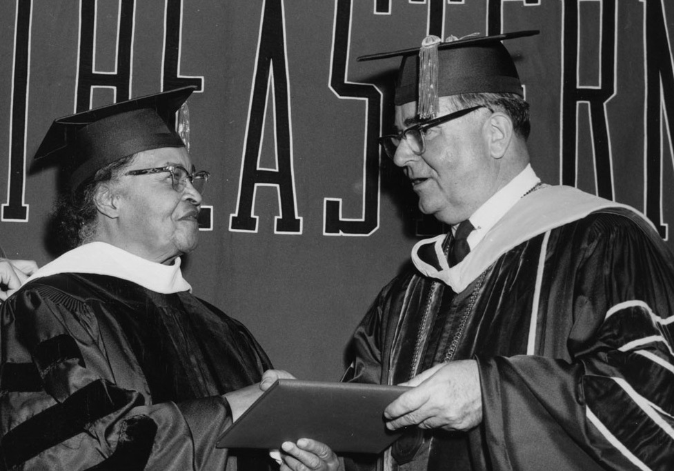 A man hands a diploma to Melnea Cass, while they shake hands. They are both wearing graduation robes and caps..