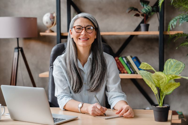 A smiling, gray haired woman seated at her desk in front of a laptop with a notepad and pen in hand.