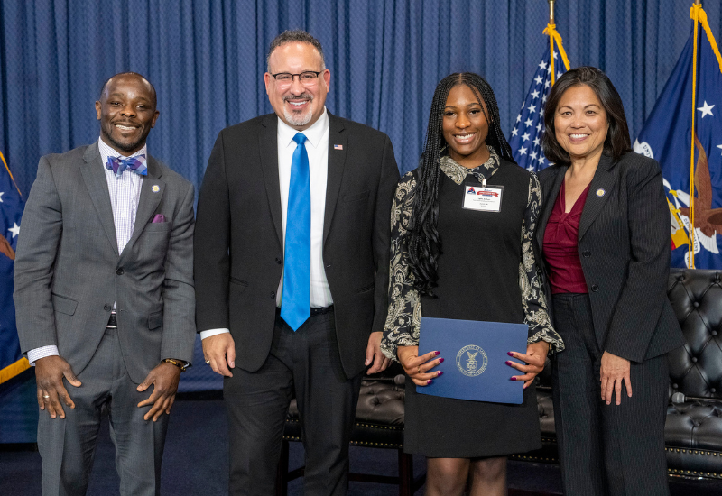 A group of four people, two men and two women wearing professional business attire, standing together and smiling for a picture in front of a blue curtain and the American flag.