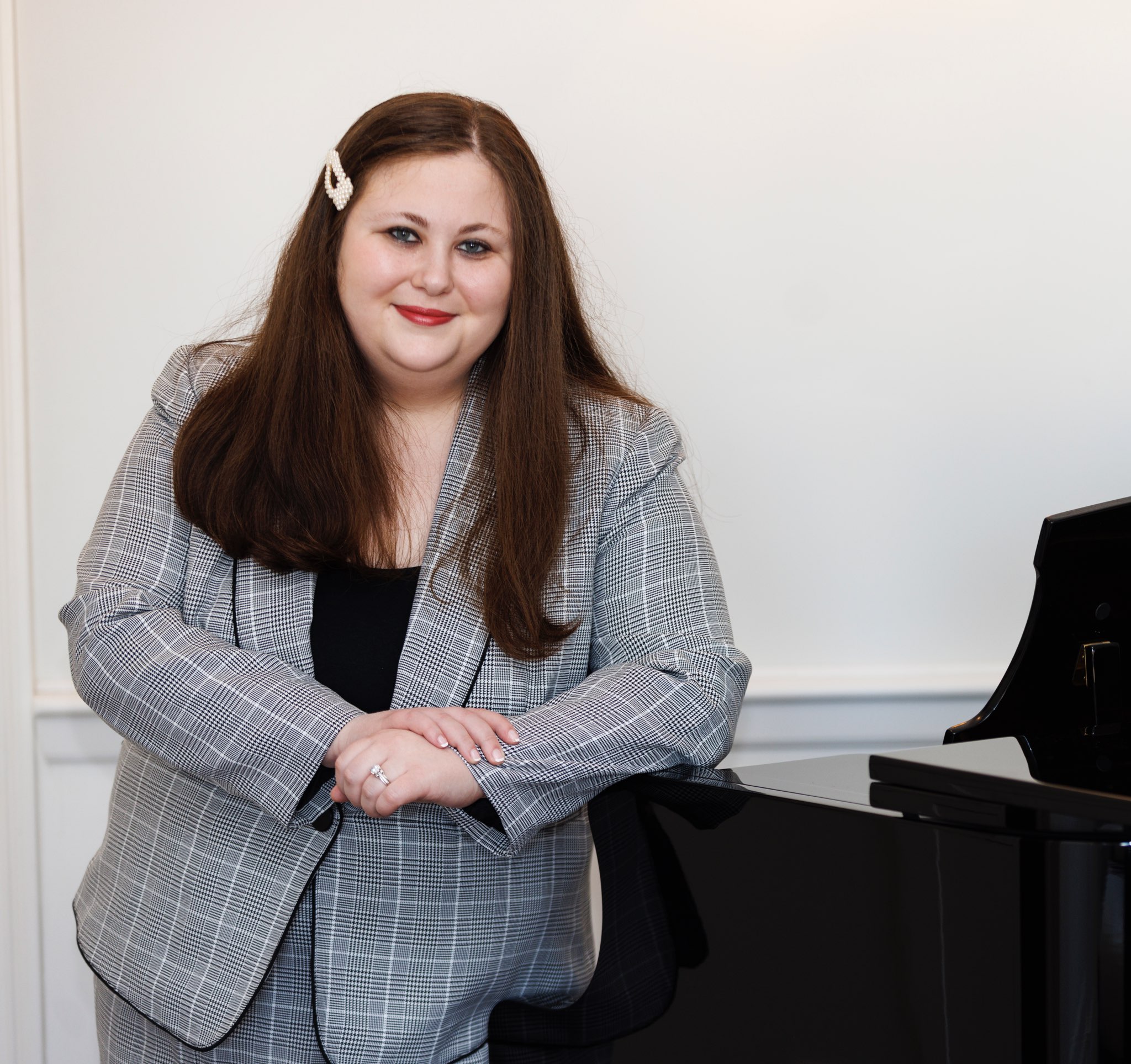 Marissa Ditkowsky - A white woman with long, dark hair in a patterned skirt suit leans on a grand piano, smiling.