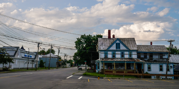 A quiet street scene in a small rural town showing an older two-story house with weathered blue and white paint under a partly cloudy sky, with electrical lines crisscrossing above.