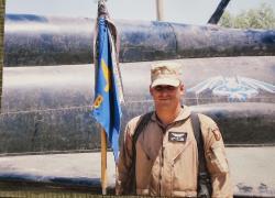 A military veteran in combat gear stands next to a military plane