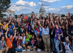 Dozens of young people gather for a rooftop group photo in front of the Capitol Building, raising their hands to the sky. 