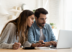 A man and woman type on a laptop. A calculator is on the table nearby.