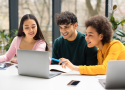 Tres jóvenes trabajan con una computadora portátil y cuadernos en una mesa, sonrientes y concentrados en su tarea. El escenario parece ser una habitación luminosa con grandes ventanales.