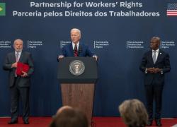 President Biden stands at a podium with Brazilian President Lula and ILO Director-General Houngbo by his side, the words "Partnership for Workers' Rights" written in English and in Portuguese above him.