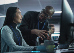 A woman sits at a computer station, engaged in conversation with a man standing next to her.