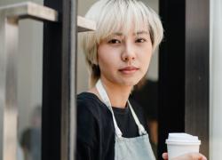 An Asian woman with platinum blond hair holding a cup of coffee wearing a black t-shirt and an apron.