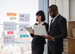 A woman and a man wearing suits holding a laptop and tablet standing by a window with sheets of paper showing graphs taped on it.