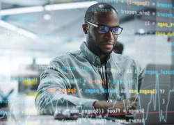 A man works on a laptop and is surrounded by computer code