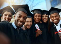 Photo of six graduates taking a selfie while dressed in their black graduation robes + hats.