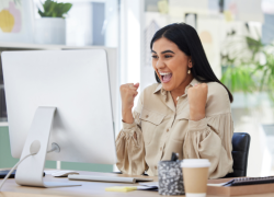 A woman with long, dark hair sits at a desk and celebrates success with raised fists and a joyful expression in front of a computer screen.