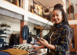 A woman holding a tablet folds sweaters on a table in a shop.