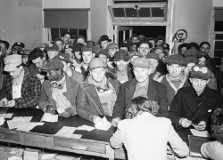 Black and white 1940s photo showing a crowd of men in work clothes clustered inside an office. There are so many that they are spilling out the door.