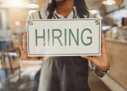 A female worker in a coffee shop wearing an apron hangs a "hiring" sign on the window.