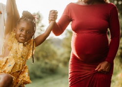 A Black family holds hands: a dad, a young daughter and a pregnant mother.