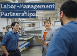 A supervisor holding a clipboard speaks with two workers in a factory environment. They all wear safety goggles and ear plugs. The text reads "Labor-Management Partnerships."