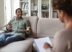 A black man with short cut hair, a beard and goatee seated on a couch and speaking with a Black woman doctor writing on a clipboard.