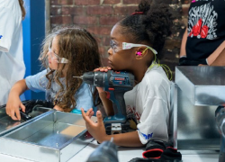 A young camper listens to an adult speak, her chin resting on a power drill, at Tools & Tiaras.