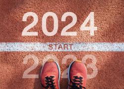 A person wearing running shoes looks down at their feet. They stand on a starting line between 2023 and 2024 written on the ground.