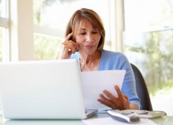 An older woman sitting at a table looking over documents with a laptop, phone and calculator laying on the table.