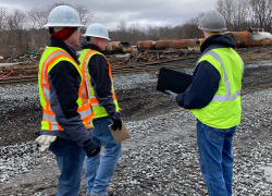 Three OSHA employees wearing hardhats and visibility vests survey the scene of the derailment.