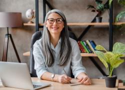 A smiling, gray haired woman seated at her desk in front of a laptop with a notepad and pen in hand.