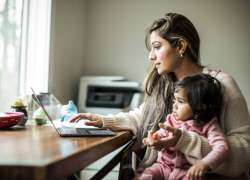 A woman types on a laptop in a home office while holding a small child. 