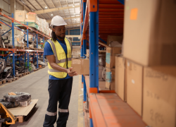 A worker wearing a hardhat and high visibility vest places a package on a shelf in a warehouse.