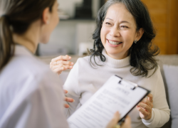 An Asian woman with dark, shoulder length hair and wearing a cream-colored turtleneck sweater smiling and looking at her doctor who is a woman with dark hair styled in a low ponytail while wearing a white coat and holding a clipboard.