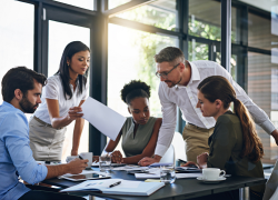 A diverse group of employees stands around a table reviewing documents.