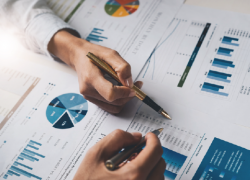 Close-up of hands holding pens above printouts of pie charts, bar charts and other data graphs. 