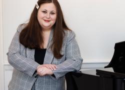 Marissa Ditkowsky - A white woman with long, dark hair in a patterned skirt suit leans on a grand piano, smiling.