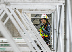 A woman in a hardhat and protective gear stands on scaffolding, overlooking a construction site.