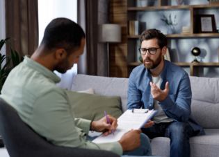 A man with fair skin and dark hair wearing glasses while seated on a couch speaking with a therapist that is wearing a green button shirt and writing on a clipboard.