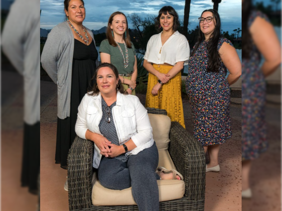 A group of five people pose for a photo outdoors at sunset. One person is sitting in a chair and the rest are standing.