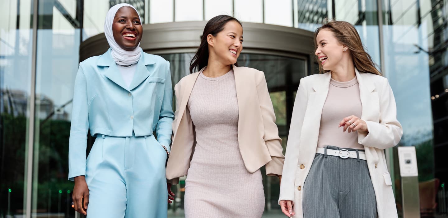 Three smiling businesswomen walk out of a building on to the sidewalk