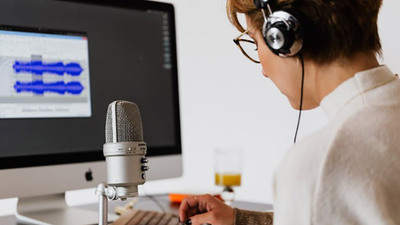 A person in headphones sits in front of a microphone tweaks a knob on a control board beneath the microphone.