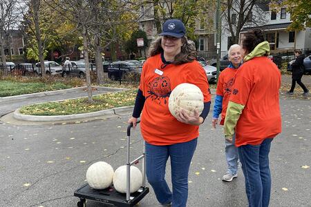 Woman wearing an orange shirt carries a white pumpkin and pulls a cart with more pumpkins