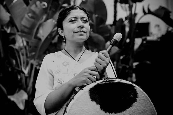 A person holds a wooden drum beater and an animal-skin drum, smiling slightly. Black-and-white photo.