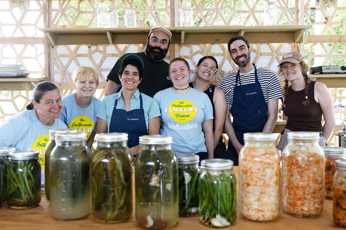 A group of people, some in pale blue Folklife Festival Volunteer T-shirts, pose behind a row of jarred pickled vegetables.